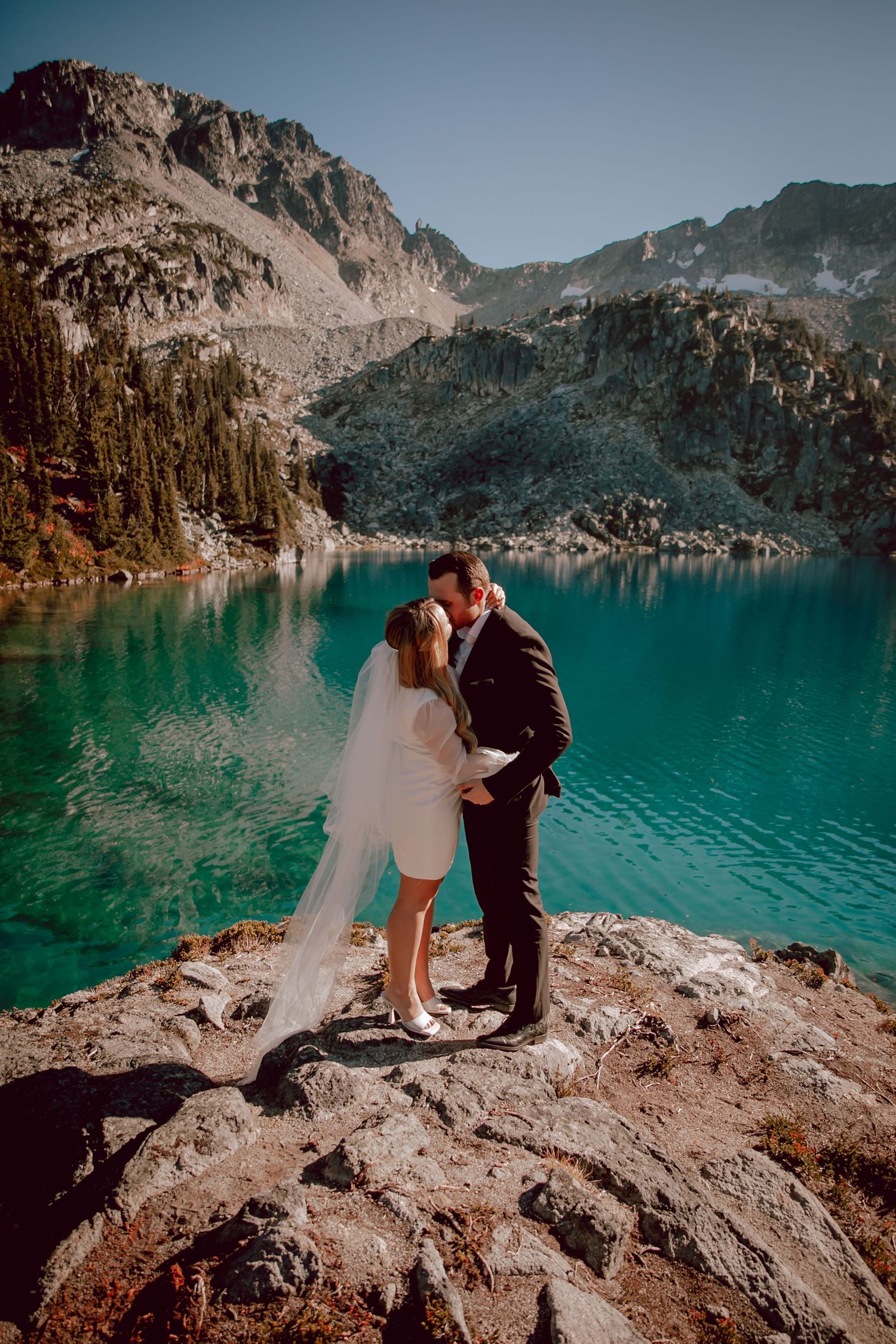 Newlyweds embrace on cliff overlooking turquoise alpine lake surrounded by mountains.