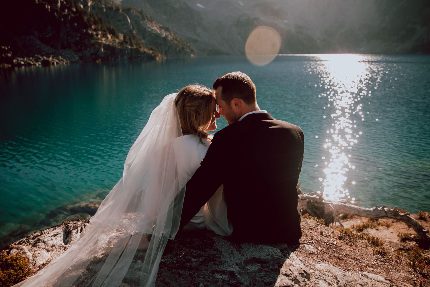 Newlyweds share intimate moment overlooking turquoise mountain lake at sunset.