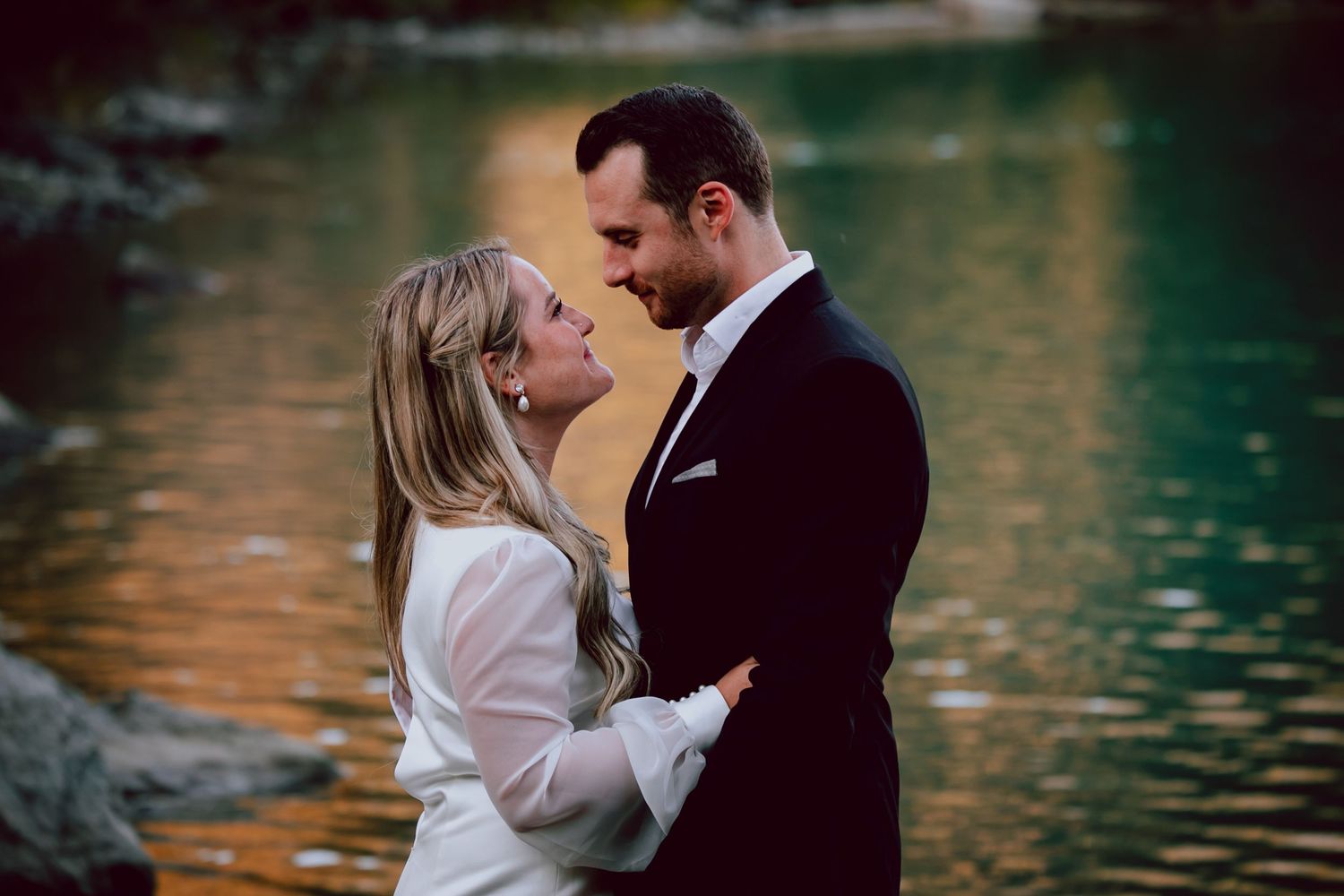 Romantic autumn wedding portrait by water with warm sunset lighting.