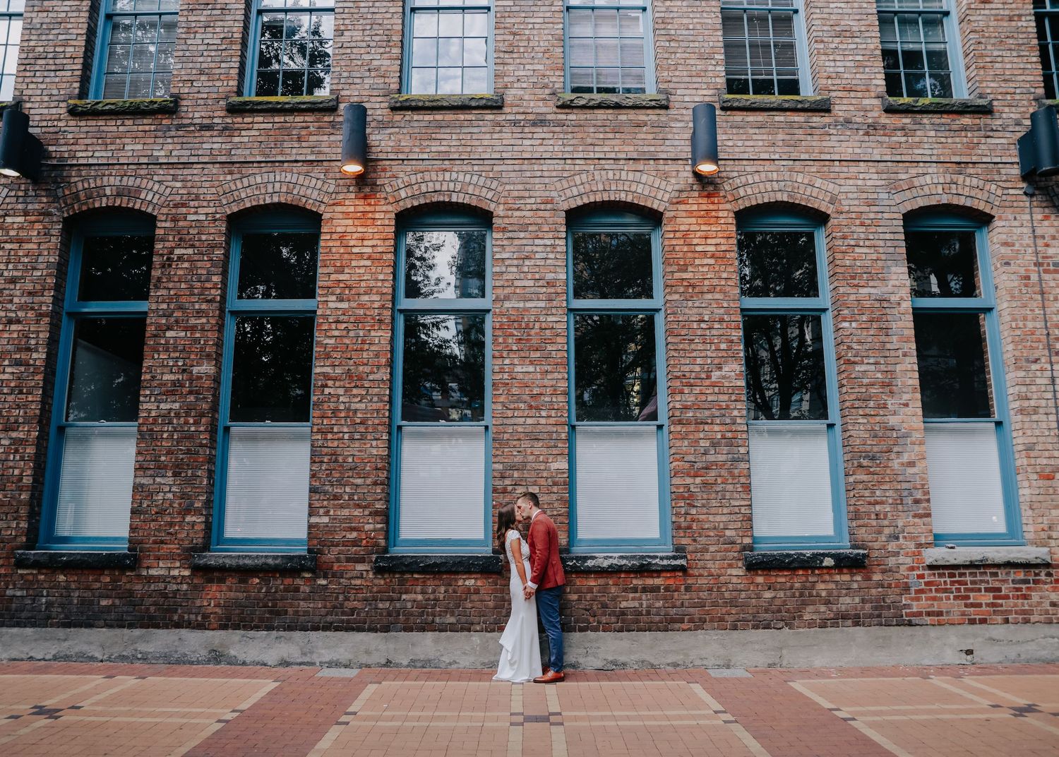 Wedding couple poses against rustic brick building with tall windows in urban setting.