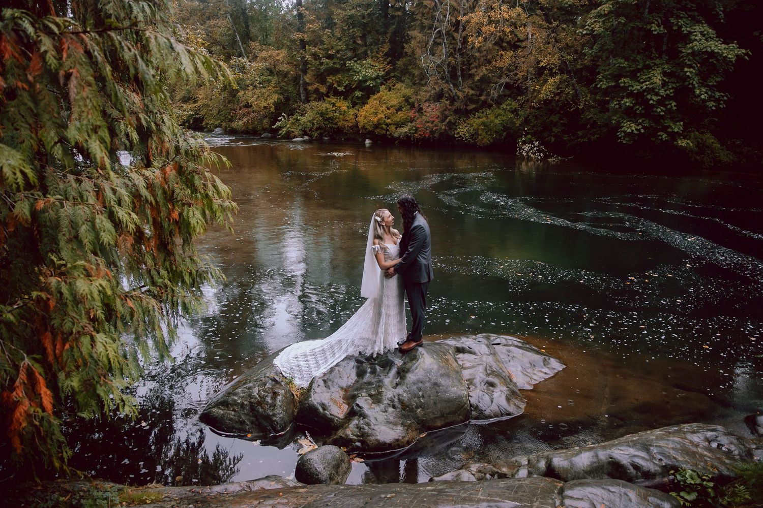 Romantic wedding portrait of couple standing on rocks by reflective forest stream.