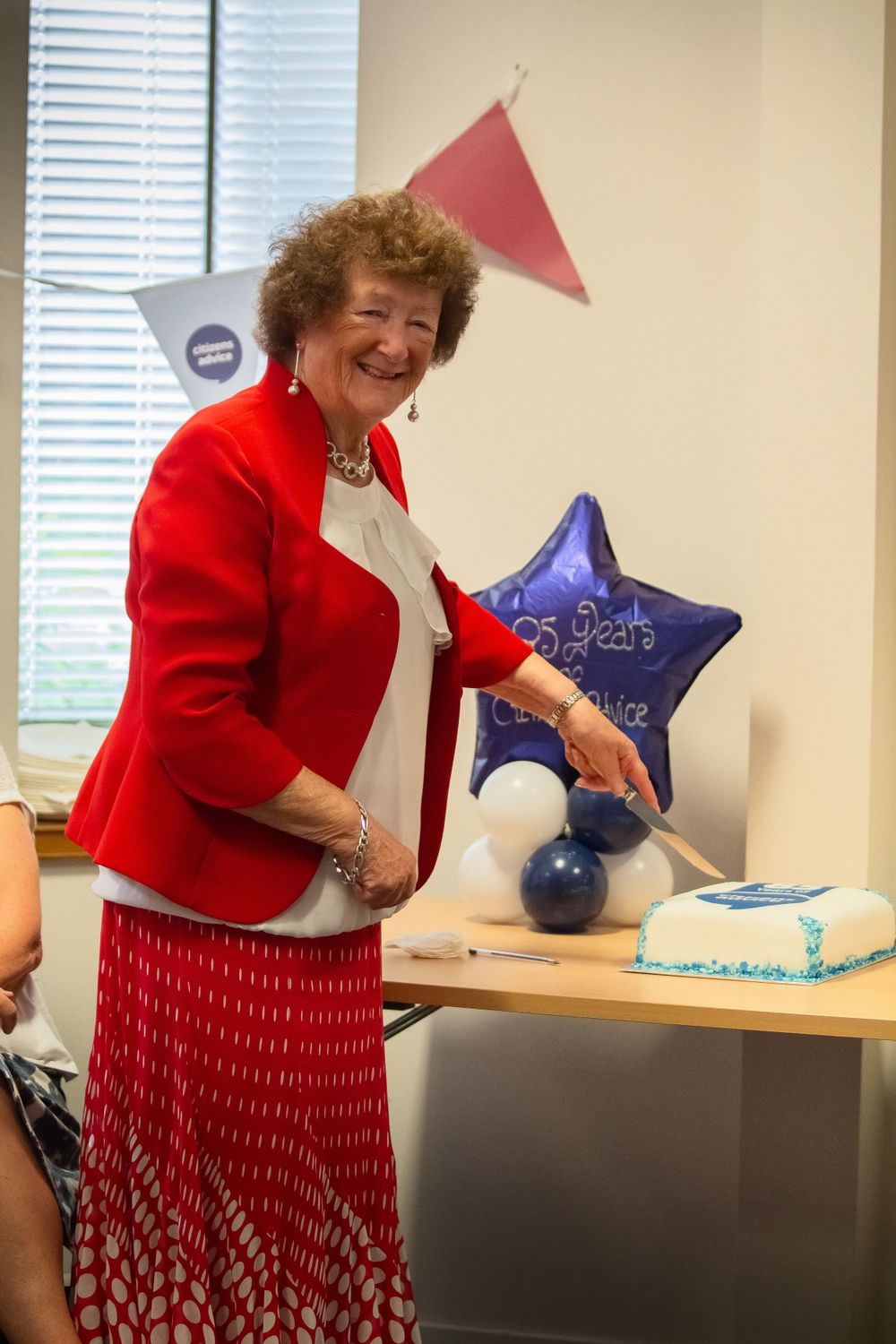 Someone in a vibrant red blazer and patterned skirt celebrates at a party with blue star balloons and a cake on the table.