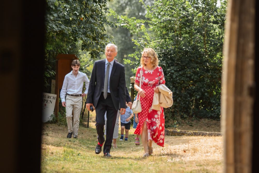 People walking along a church pathway on a sunny day, with a woman in a flowy red dress and a man in a suit.