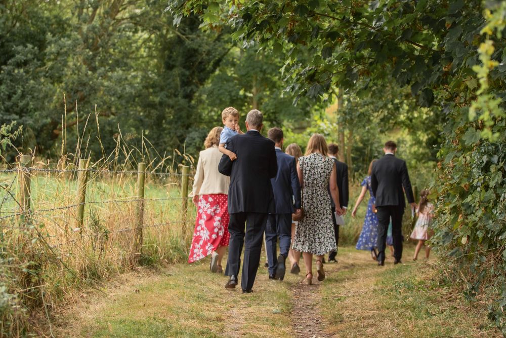 A group of formally dressed people walk together along a scenic countryside path on a sunny summer day.