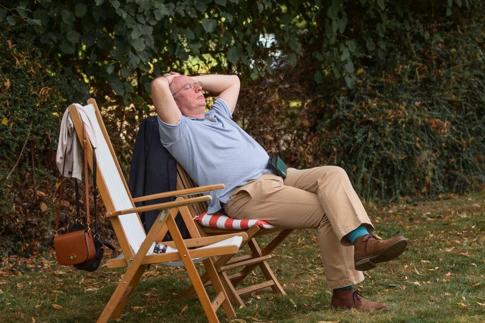 Person relaxing in a wooden deck chair on grass with hands behind head in a peaceful garden setting.