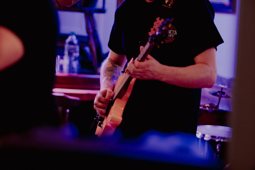 Musicians perform on stage with electric guitars in a dark venue with purple lighting effects.