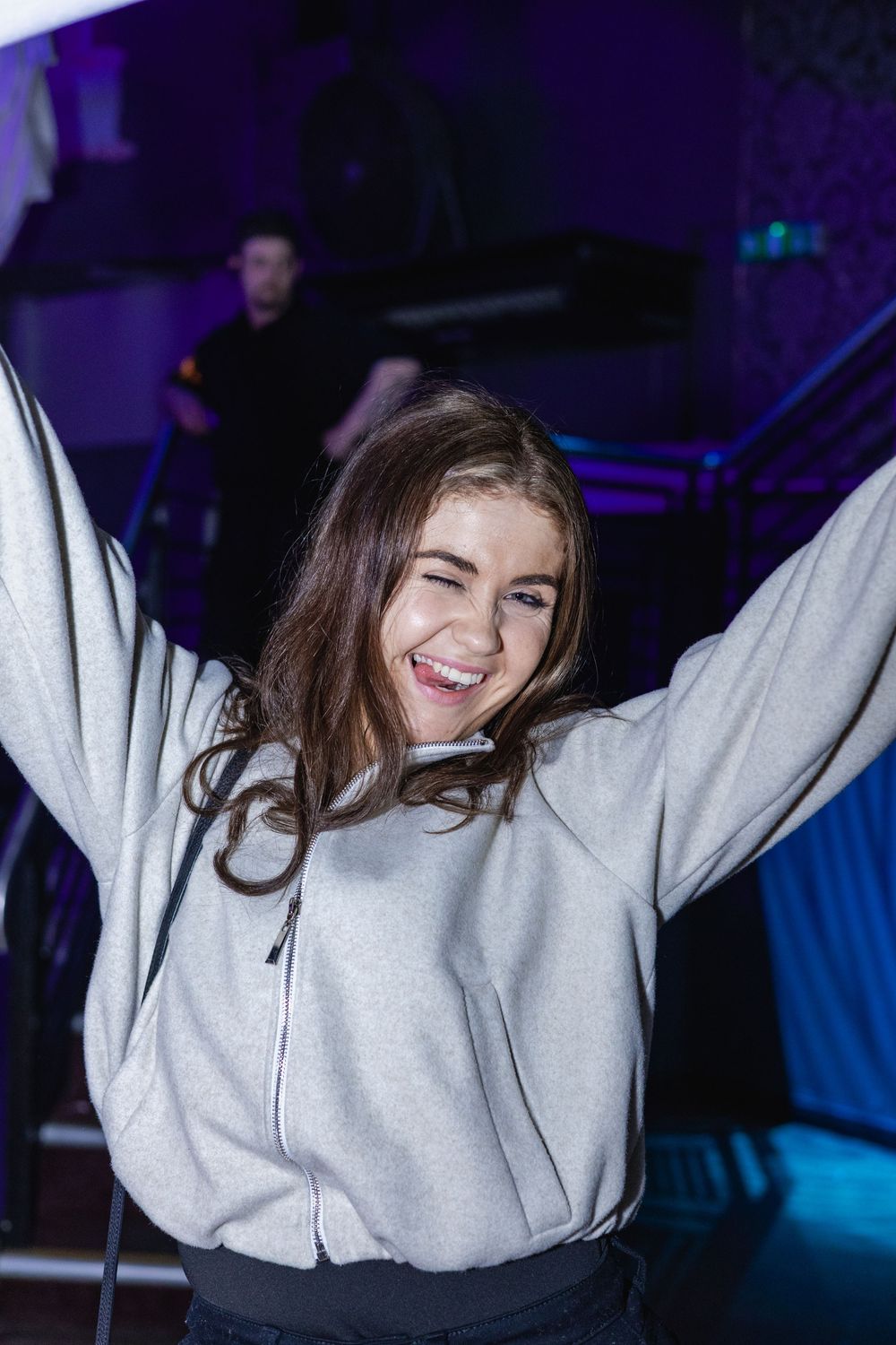 Person in light gray sweatshirt dancing with raised arms against dark background with blue lighting.