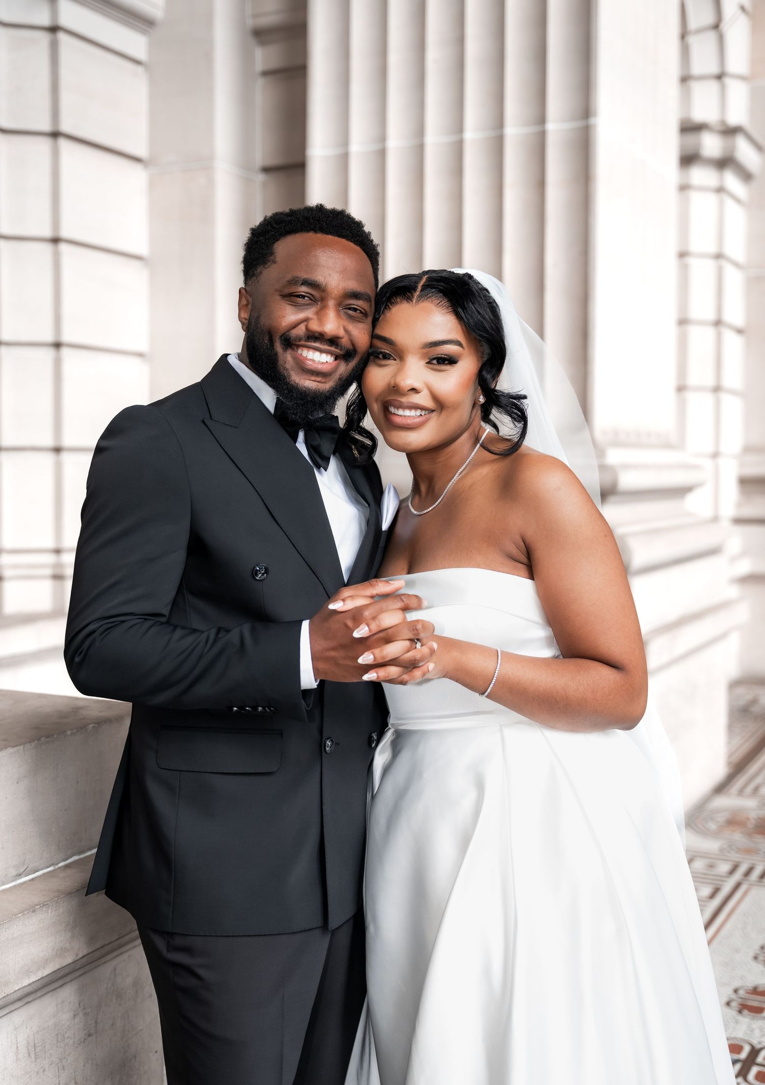 A happy newlywed couple poses together in formal wedding attire against classic white columns of an elegant building.