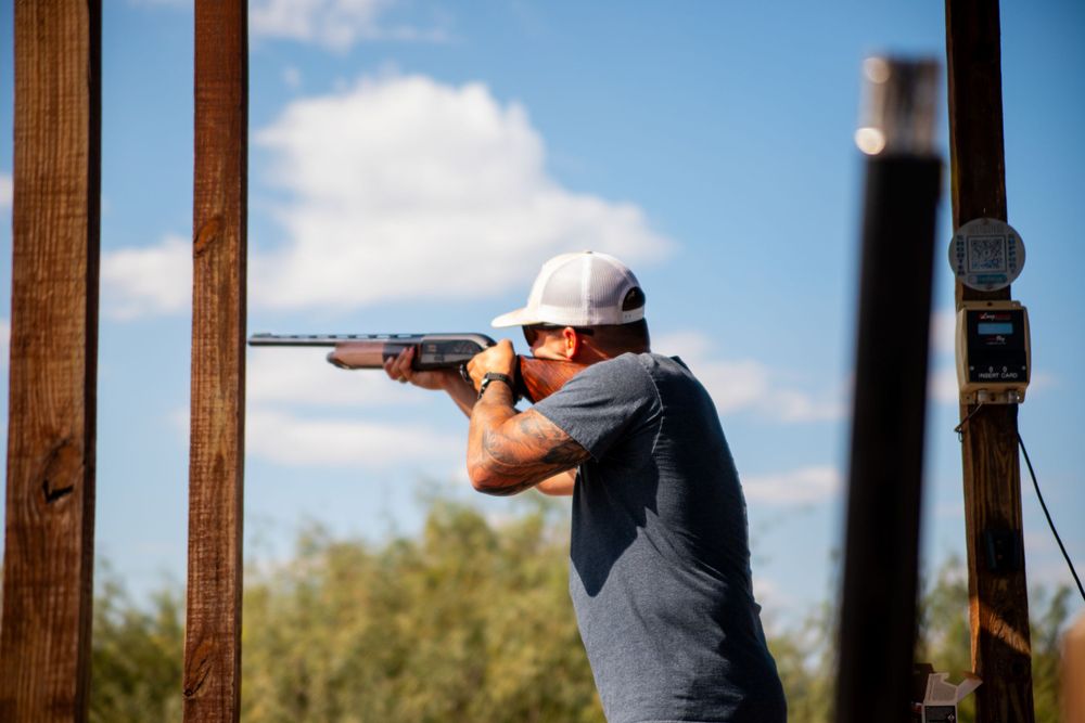 Person in gray shirt aiming a shotgun at clay target shooting range.