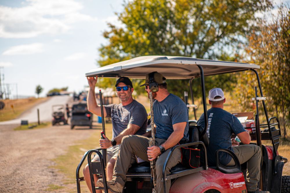 People riding in golf cart on dirt path at outdoor sporting venue.