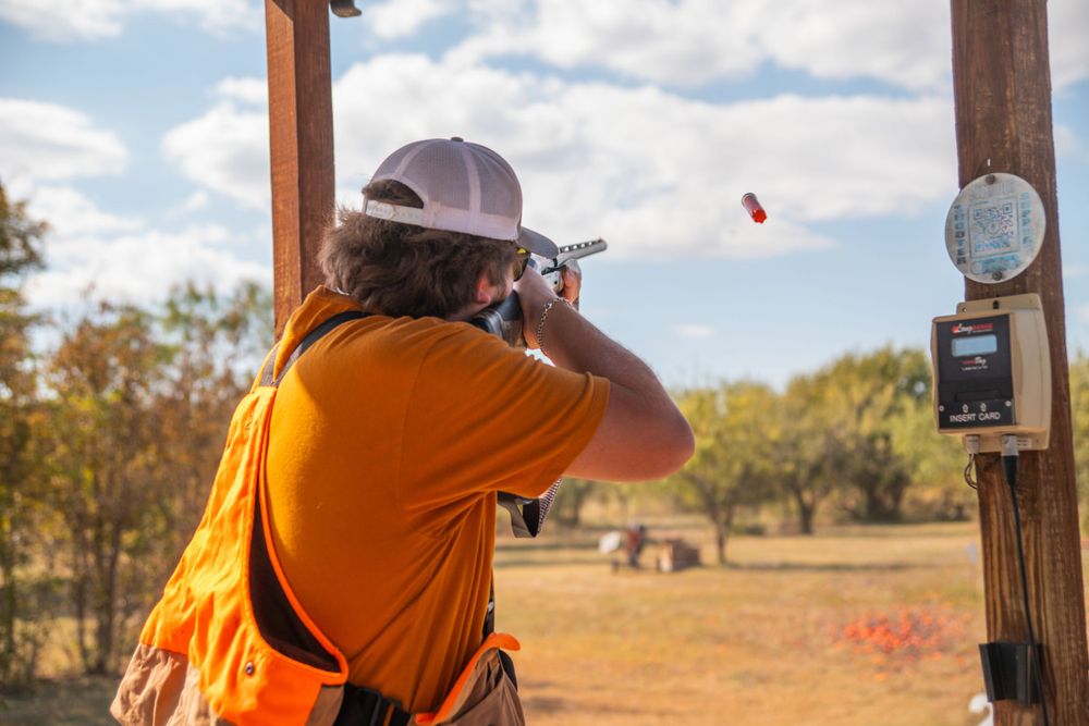 Person in orange safety vest shooting at clay targets on outdoor range.