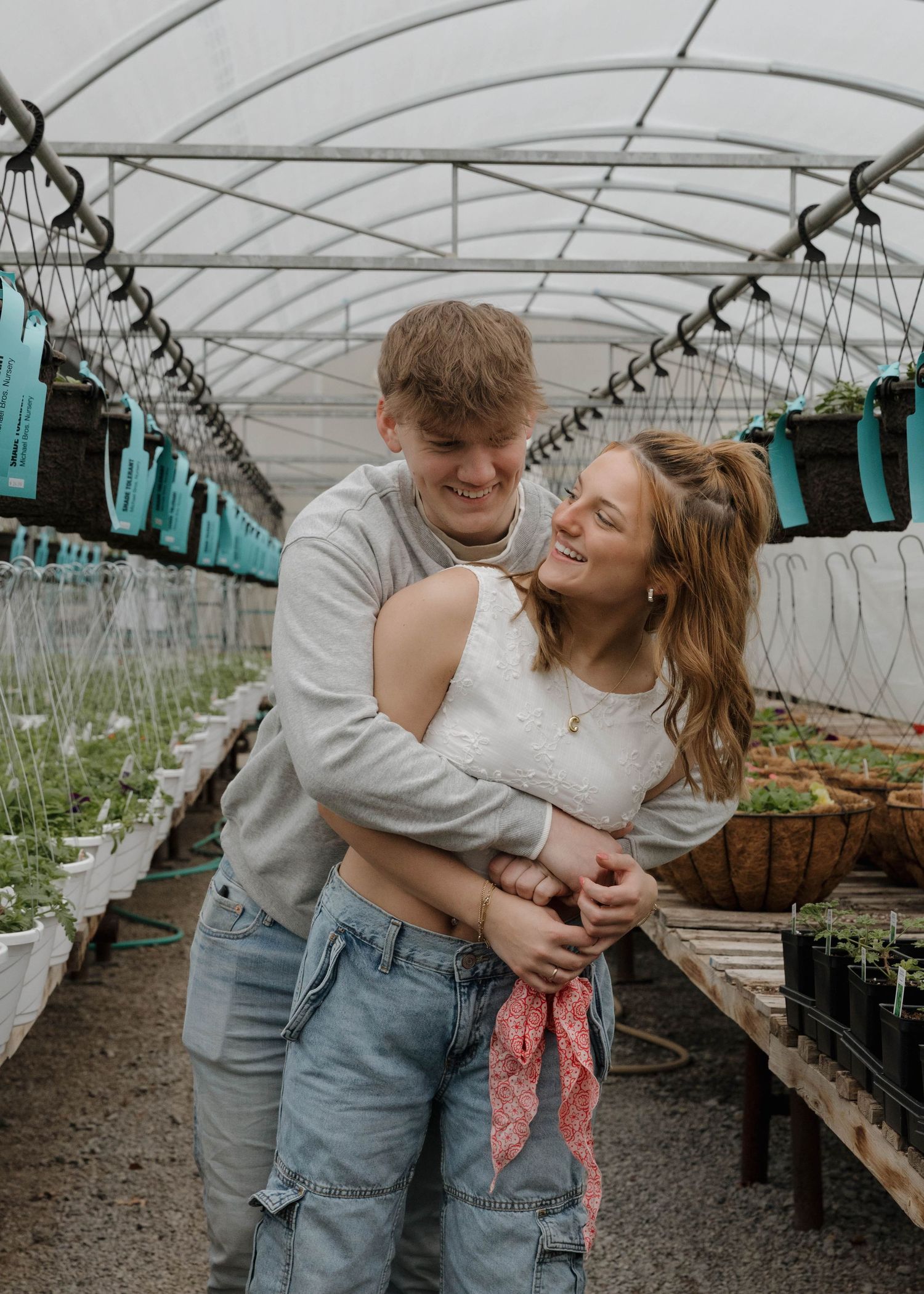 A couple embraces and laughs together inside a greenhouse with hanging planters and garden beds.