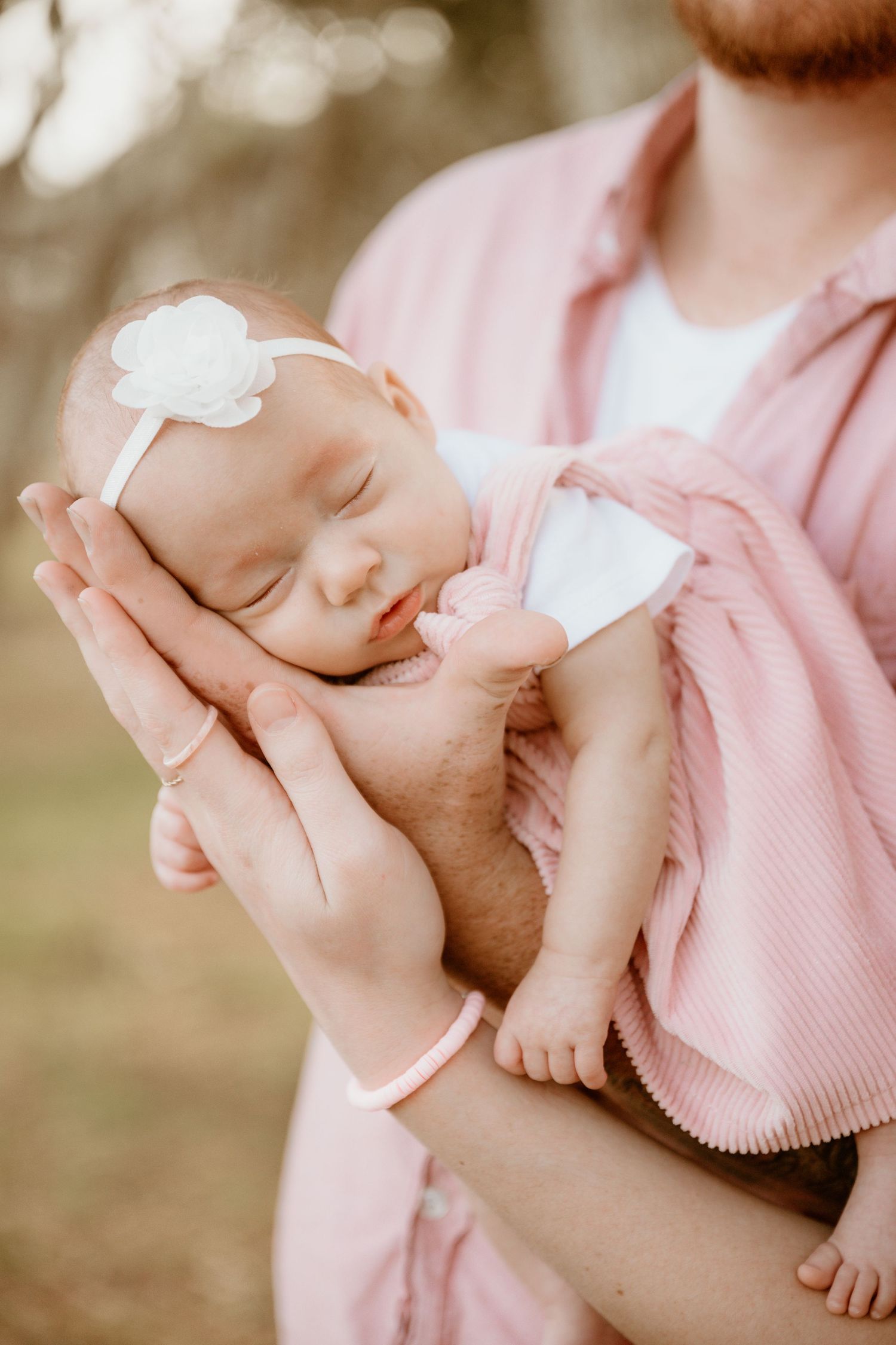 Father with red beard gently cradles sleeping newborn baby girl in pink outfit with white flower headband.