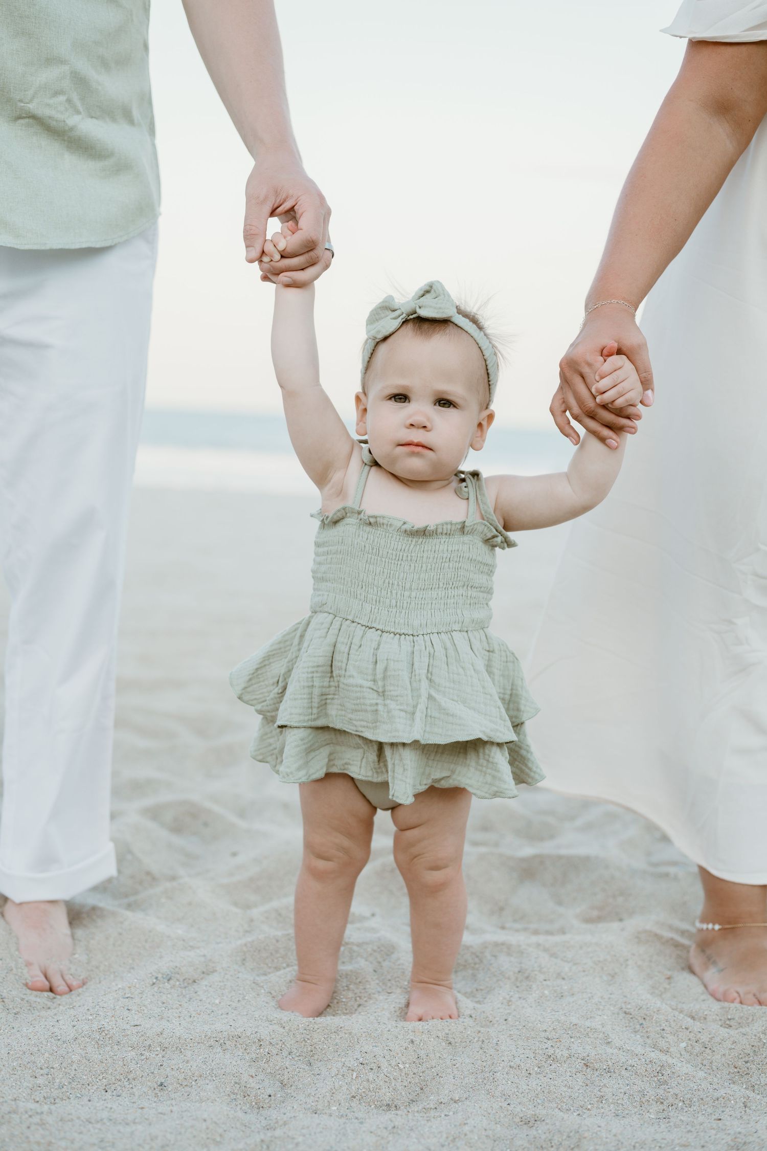 Baby girl in sage green ruffled dress holds parents hands while standing on sandy beach at sunset.