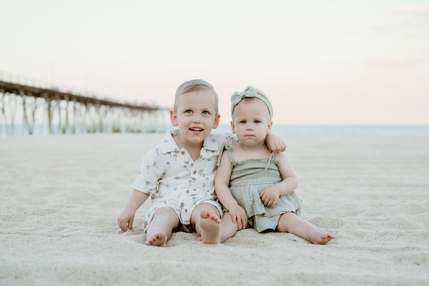 Two young siblings sit together on sandy beach, older toddler boy and baby girl in sage green outfit with bow headband.