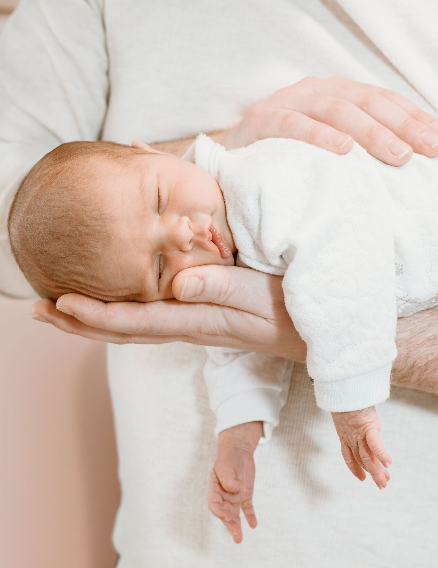 Sleeping newborn baby in white outfit rests peacefully in parent's hands against soft white background.