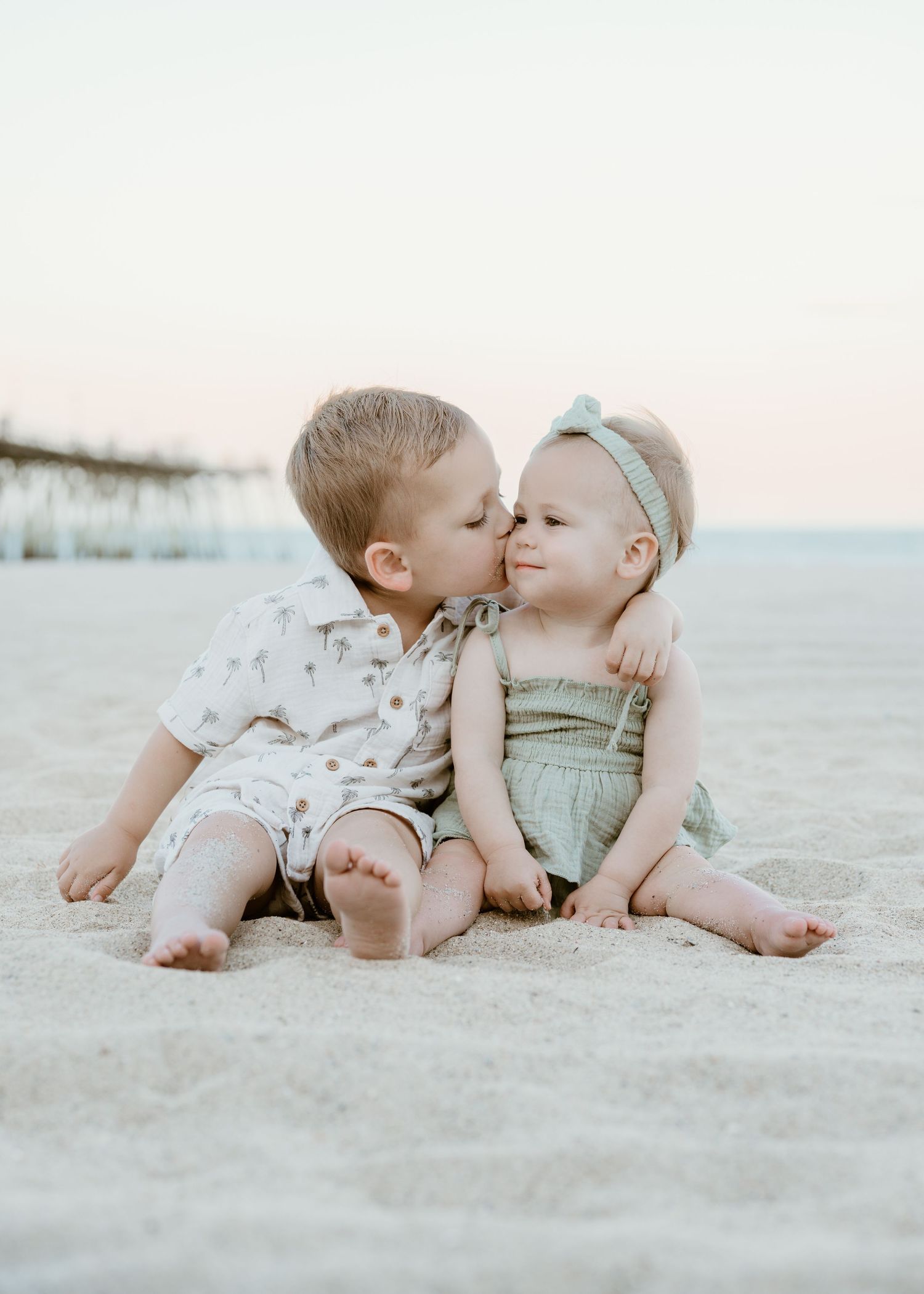 Sweet toddler boy kisses baby sister on cheek while sitting on sandy beach near pier at sunset.