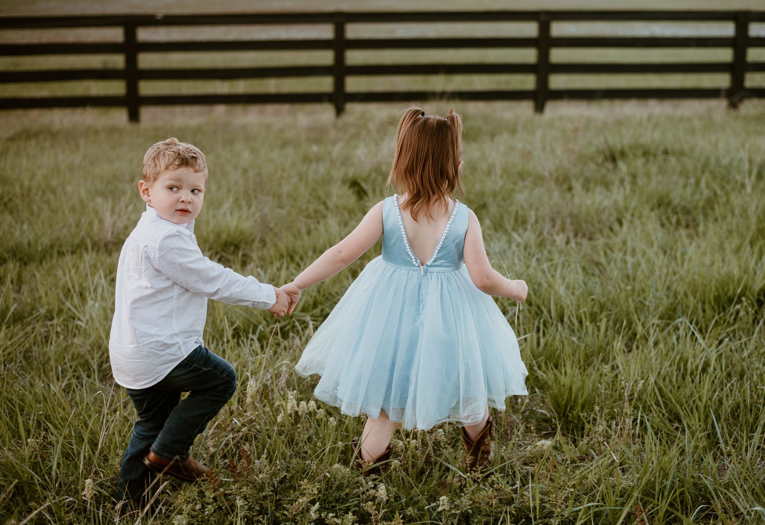 Two children hold hands while running through a grassy field, with one wearing a blue tulle dress.