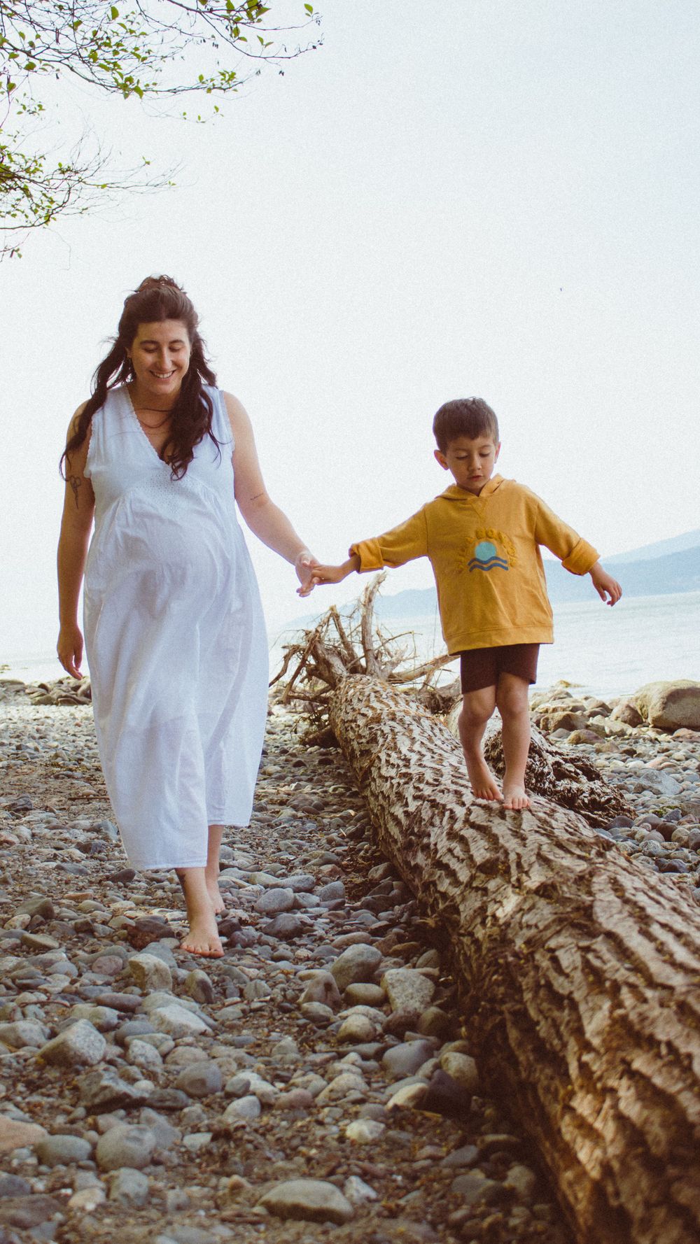 pregnant mom in white flowy dress holding hands with toddler while he's balancing on a log at the beach during a family maternity session.