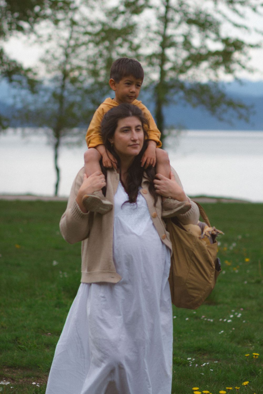 toddler sitting on mom's shoulders while she is walking through a park showing her pregnant belly during a family maternity session.