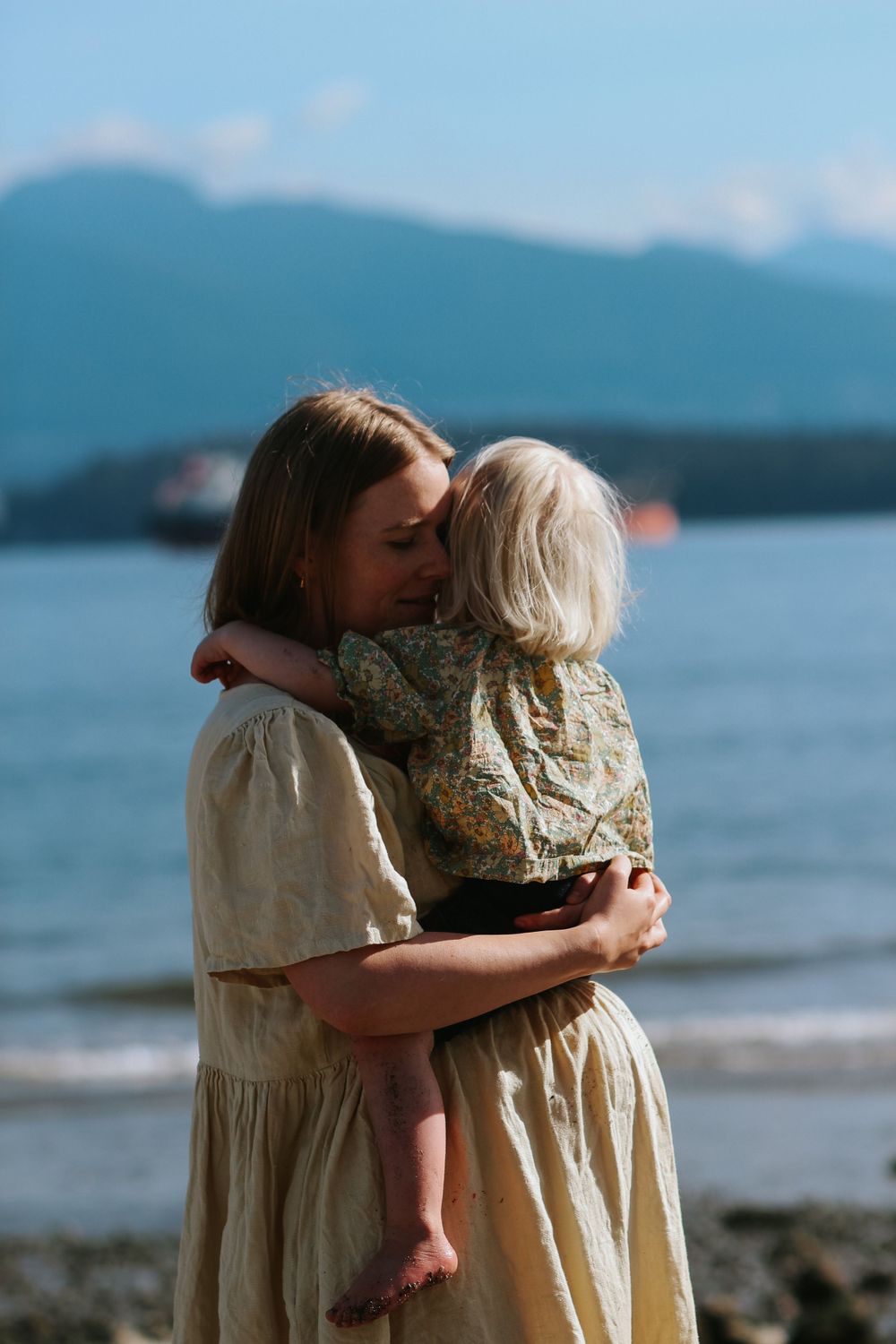 pregnant mom cuddling her blonde toddler who is sitting on her baby bump during a family maternity session on the beach in vancouver, bc.