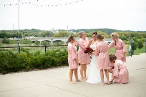 Bridesmaids in pink robes help adjust the bride's dress outdoors with string lights visible above.