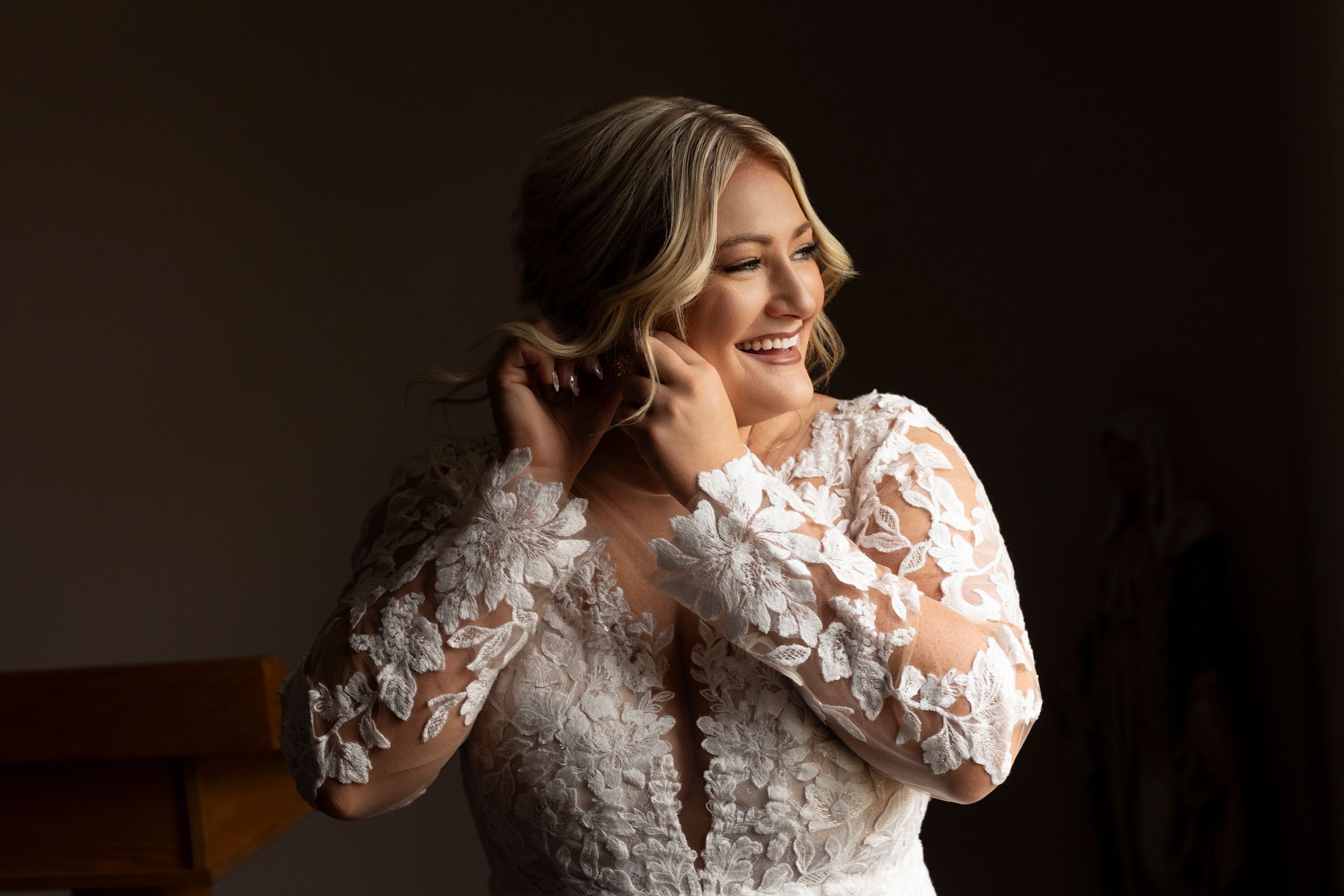 Bride in lace long sleeve dress adjusting earrings in dramatic window light.