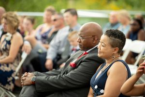 Wedding guests seated outdoors watching ceremony on white chairs during daytime event.