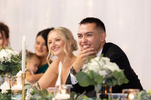 Newlyweds and wedding party guests smile while seated at an elegantly decorated reception table with white floral centerpieces.