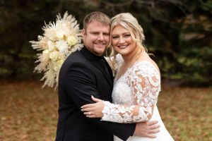 Wedding couple embraces while holding white and cream pampas grass bouquet in autumn setting.