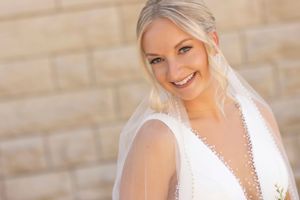 A bride in a white wedding dress and veil smiles radiantly against a stone wall background.