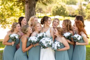 Wedding party in sage green dresses shares joyful moment with bride outdoors.