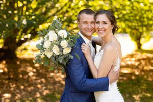 A wedding couple embraces outdoors on a sunny day with a beautiful white rose bouquet.