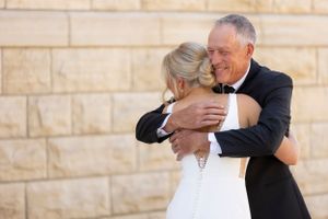 A tender moment between a bride in white and someone in a black suit embracing against a stone wall.