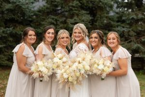 Bridesmaids in light colored dresses hold elegant cream-colored bouquets while posing against evergreen trees.