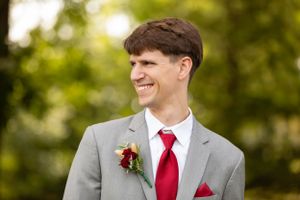 Groom in grey suit and red tie smiles while wearing boutonniere during outdoor wedding.