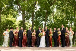 Large wedding party lined up for formal photo in burgundy and black attire under trees.