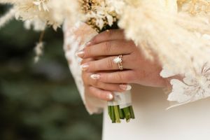 Close-up of wedding rings and hands holding a cream-colored bouquet with greenery wrapped stems.