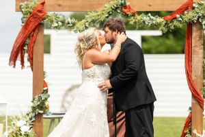 A romantic wedding ceremony takes place under a rustic wooden arch decorated with greenery and red ribbons.