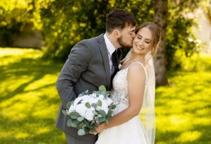 A romantic wedding portrait of a couple embracing in a sunlit garden with a white floral bouquet.