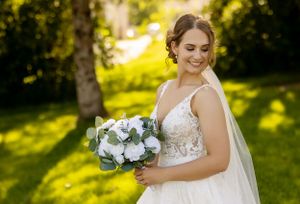 A bride in a lace wedding gown holds a white and green bouquet in a sunny outdoor setting.