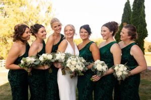 Bridesmaids in emerald green dresses hold white bouquets while laughing with the bride.