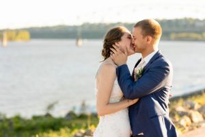A romantic wedding portrait of a couple kissing by a waterfront at sunset.