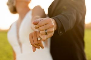 Newlyweds hold hands showing wedding rings during golden hour outdoor portrait session.