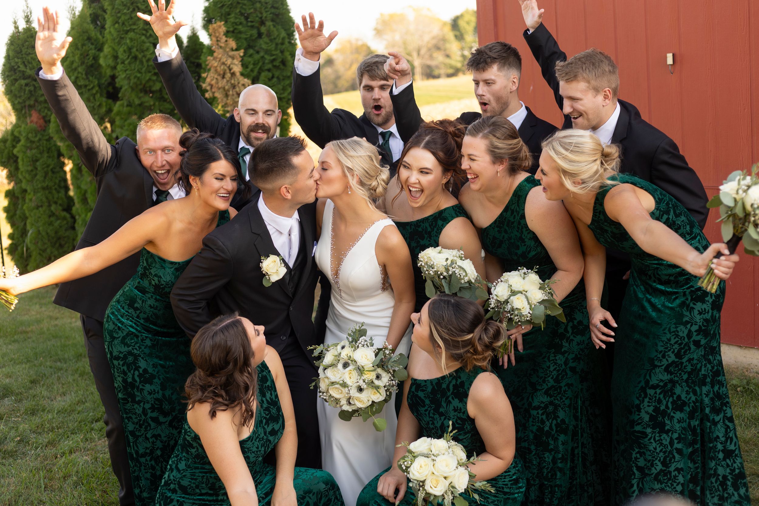 Wedding party strikes a fun, energetic pose outdoors near a red barn.