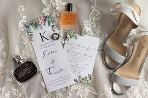 Wedding details flatlay featuring invitation, perfume bottle, and silver sandals on lace fabric.