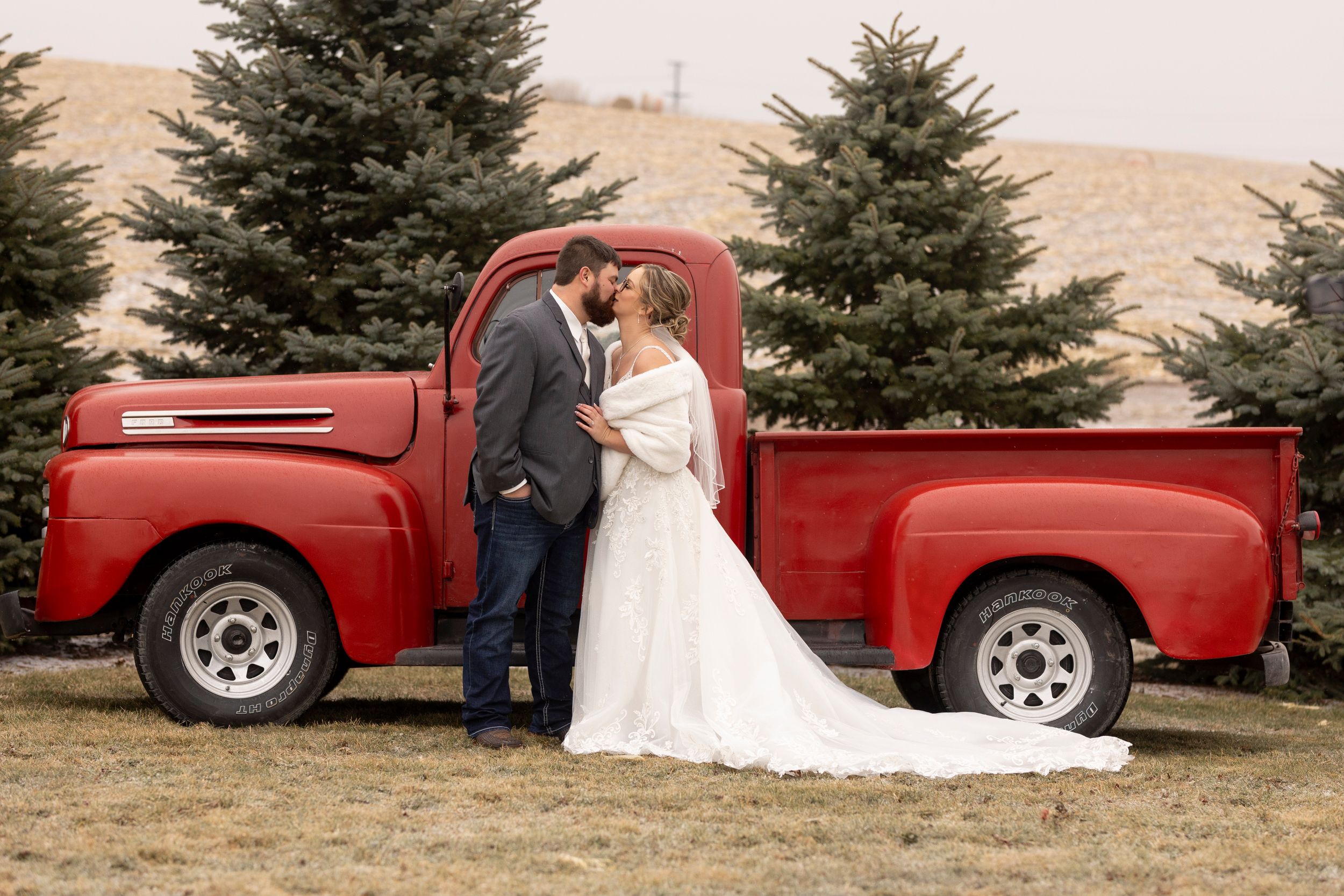 Wedding couple sharing romantic moment in front of vintage red pickup truck with evergreens in background.