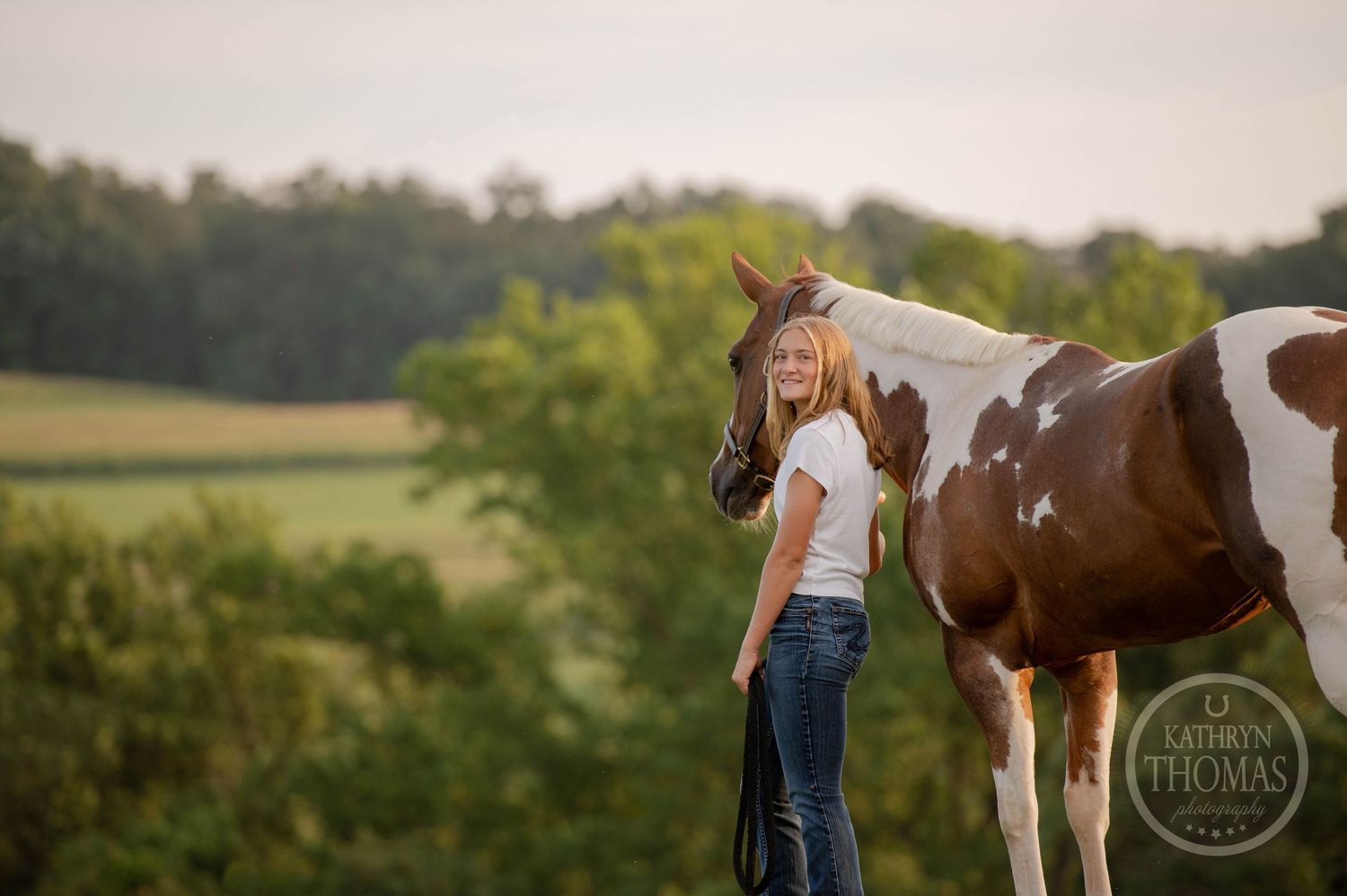 Paint horse and person stand together in peaceful countryside setting during golden hour.