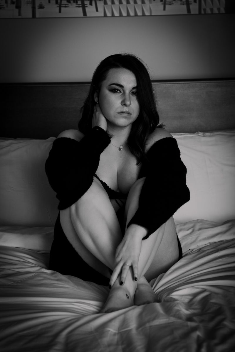 Intimate black and white boudoir photo of a person in a dark cardigan posing on white bedding.