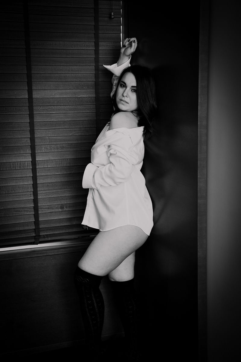 Black and white boudoir photo of someone posing in an oversized shirt against a window with blinds.